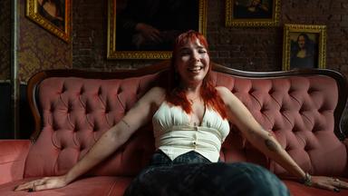Eine junge Frau sitzt mit ausgebreiteten Armen und offenen Beinen, Top und Jeans, auf einem Sofa und lacht.