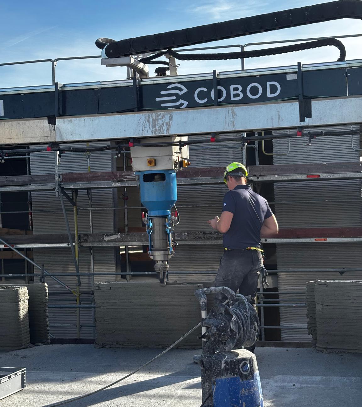Auf einer Baustelle beaufsichtigt ein Bauarbeiter mit Schutzhelm einen 3D-Drucker, der einen Spezialzement aufträgt.