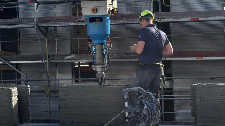 Auf einer Baustelle beaufsichtigt ein Bauarbeiter mit Schutzhelm einen 3D-Drucker, der einen Spezialzement aufträgt.