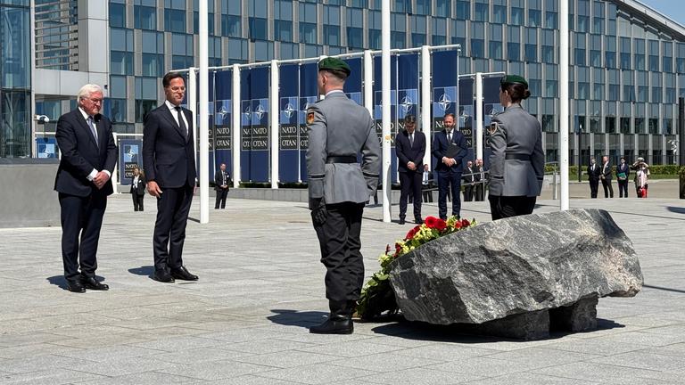 Steinmeier und Rutte bei Festakt in Brüssel. Militärangehörige stehen ihnen gegenüber, zwischen ihnen ein Blumenkranz