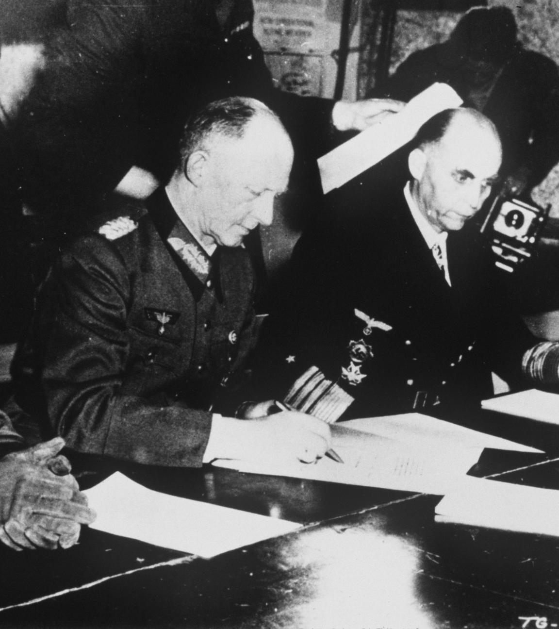 Major Wilhelm Oxenius, Generaloberst Alfred Jodl und Generaladmiral Hans-Georg von Friedeburg sitzen an einem Tisch und Jodl unterzeichnet das vor ihm liegende Dokument.