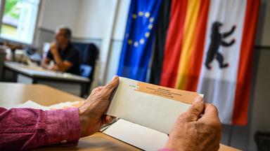 Ein Wahlhelfer hält einen Stimmzettel in einem Wahlumschlag in einem Wahllokal in der Hand. Im Hintergrund die Flagen der EU, Deutschlands und Berlins.
