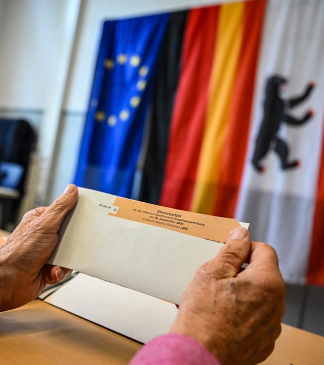 Ein Wahlhelfer hält einen Stimmzettel in einem Wahlumschlag in einem Wahllokal in der Hand. Im Hintergrund die Flagen der EU, Deutschlands und Berlins.