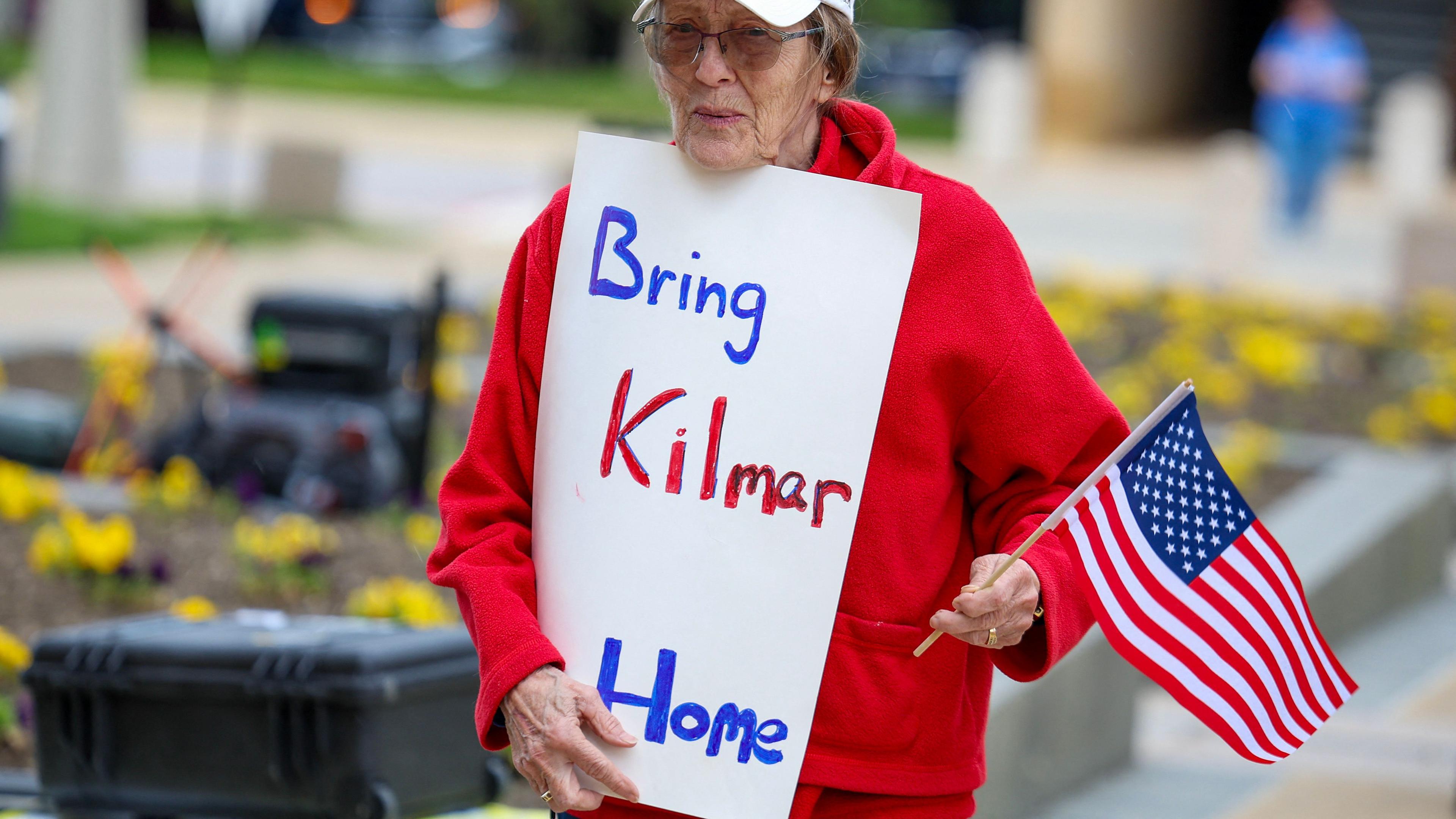 Eine Frau trägt auf einem Protest eine kleine US-Flagge und ein Schild mit der Aufschrift "Bring Kilmar Home".