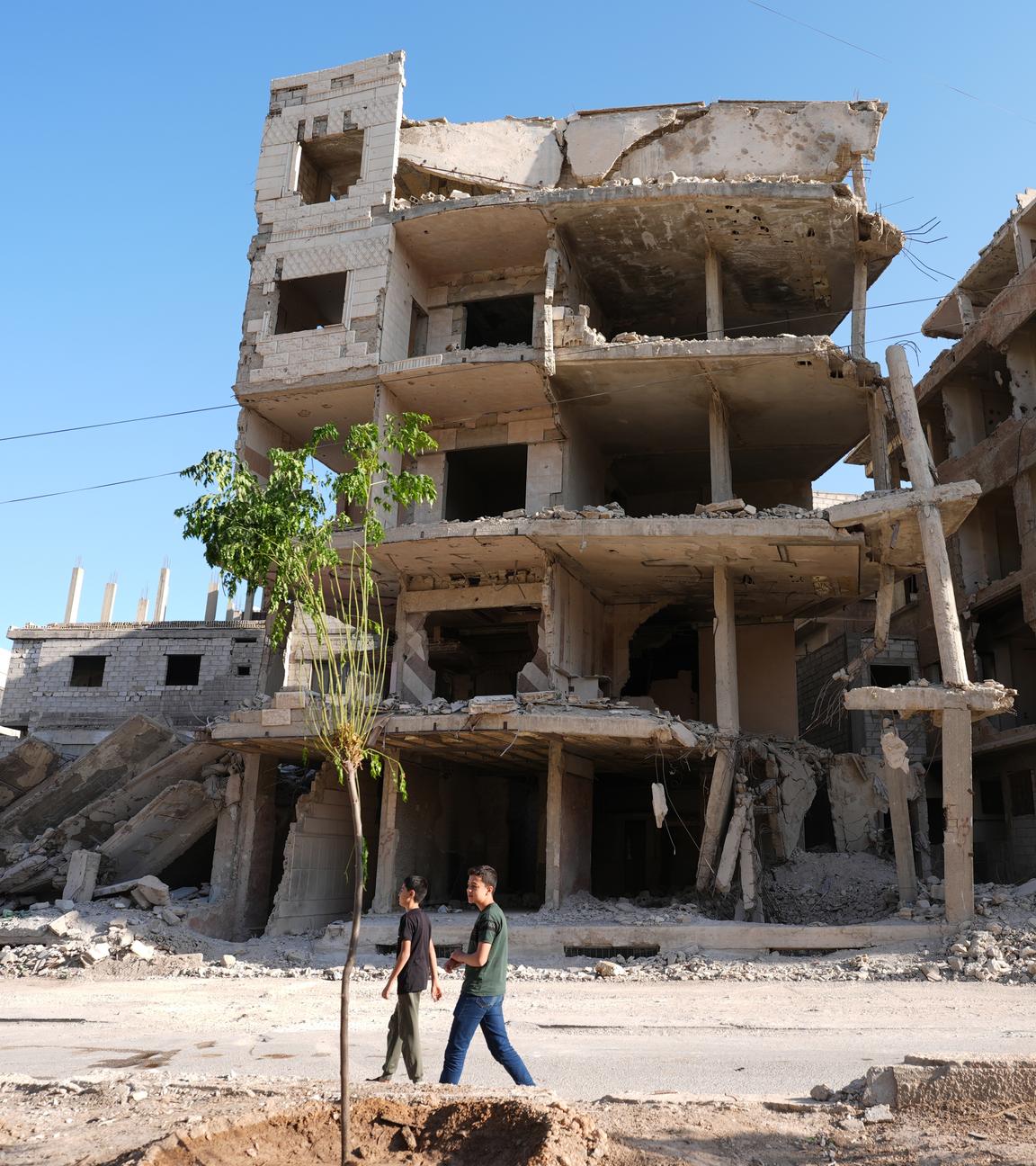 Auf dem Bild ist eine Ruine in Syrien zu sehen. Zwei Kinder laufen daran vorbei.