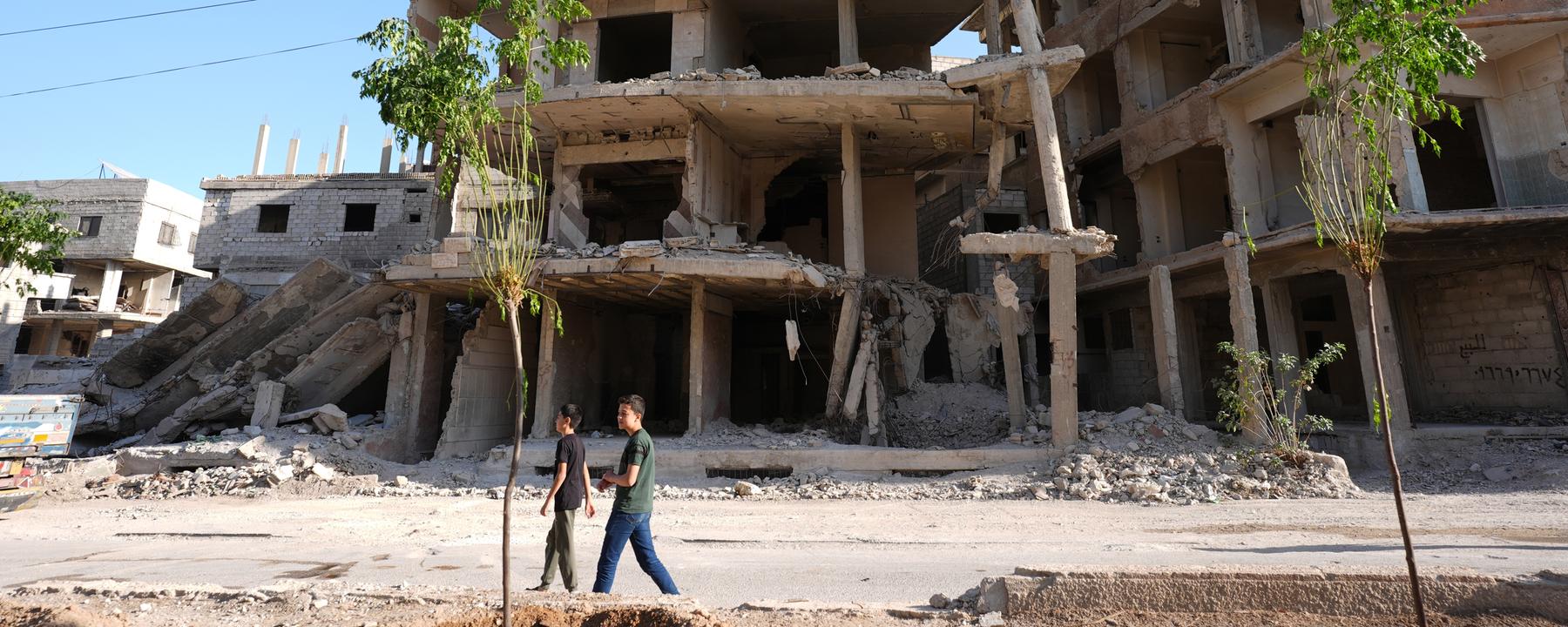 Auf dem Bild ist eine Ruine in Syrien zu sehen. Zwei Kinder laufen daran vorbei.