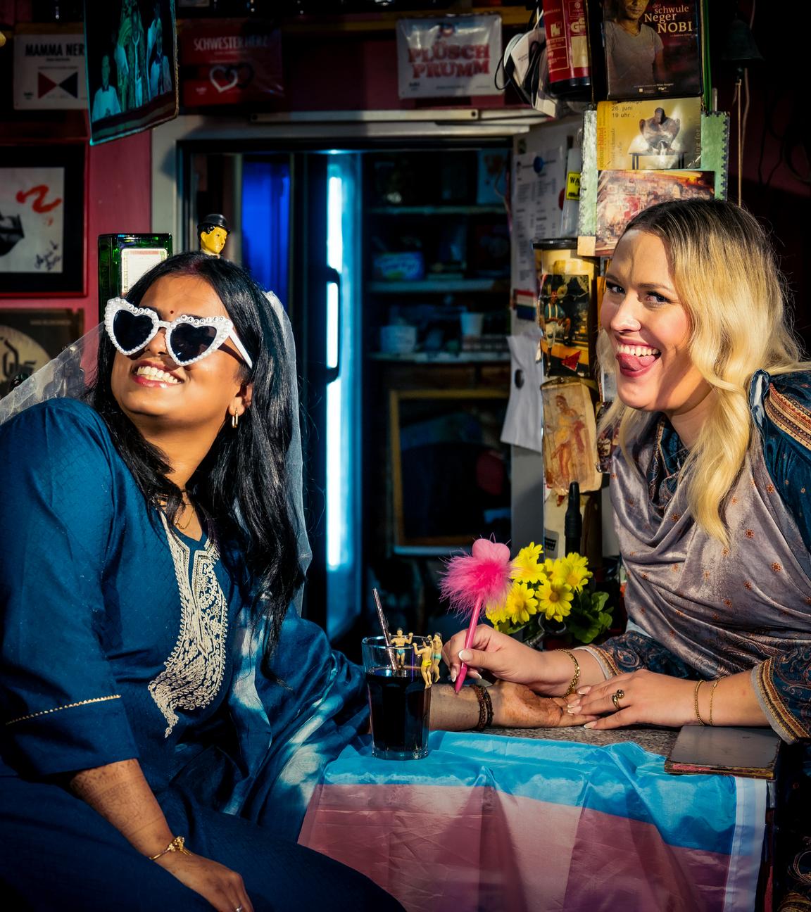 Hinduistin Sagitha blickt lächelnd seitlich nach vorne. Sie trägt eine Sonnenbrille in Herzform, auf der "just married" steht, und einen Schleier. Atheistin Josi sitzt im Sari neben ihr und streckt ihre Zunge heraus.