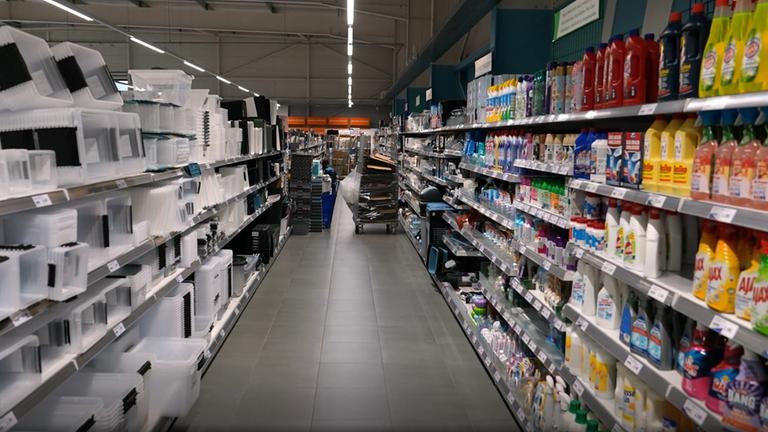 Ein Gang in einem Laden, in den Regalen unterschiedliche Produkte. Rechts stehen Plastikaufbewahrungskisten, links Reinigungsmittel.