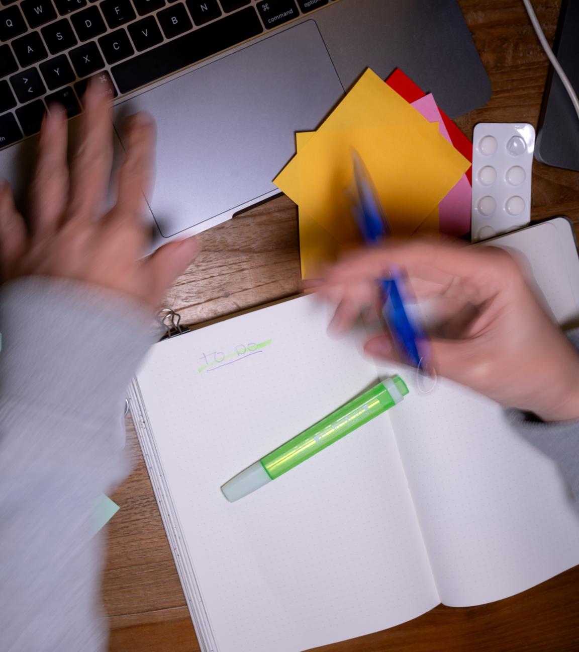 Eine Frau sitzt an einem Schreibtisch, auf dem Tabletten, Post-Its, ein Laptop, ein Tablet und ein Notizbuch liegen und erstellt eine To-do-Liste.