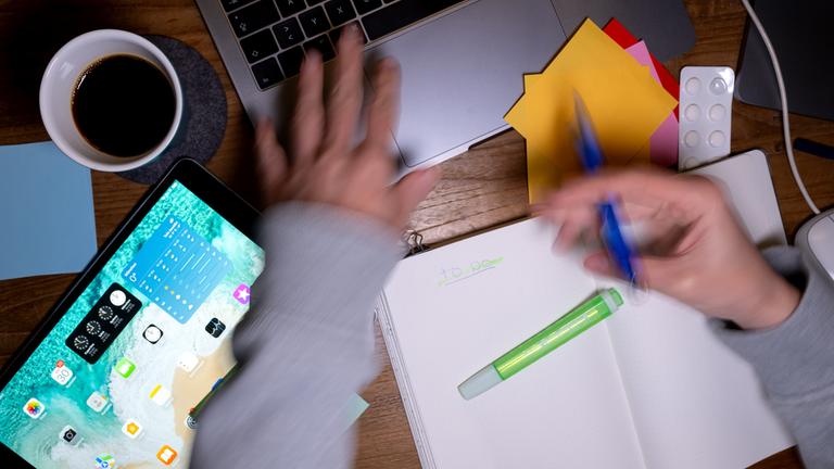 Eine Frau sitzt an einem Schreibtisch, auf dem Tabletten, Post-Its, ein Laptop, ein Tablet und ein Notizbuch liegen und erstellt eine To-do-Liste.