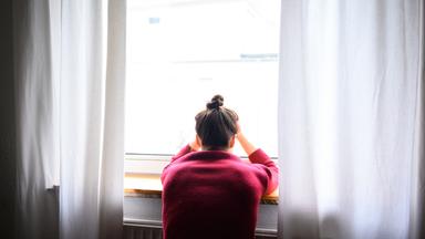 Symbolbild: Einsame Frau alleine und verzweifelt am Fenster und stützt Hände auf den Kopf