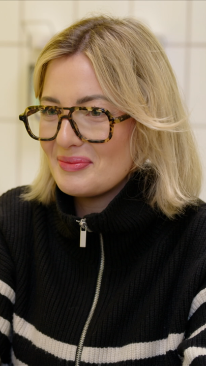 Blonde Frau mit Hornbrille mit verschmitztem Gesichtsausdruck im Gespräch.