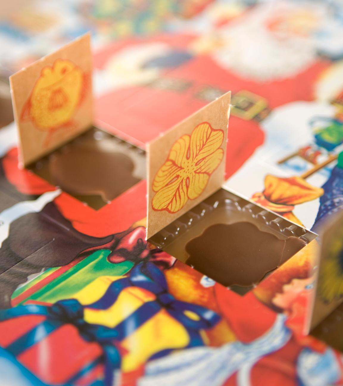 Man sieht einen Adventskalender mit winterlichem Motiv an dem vier Türchen nebeneinander geöffnet sind. Die kleinen Schokoladenmotive liegen noch in ihren Förmchen. 