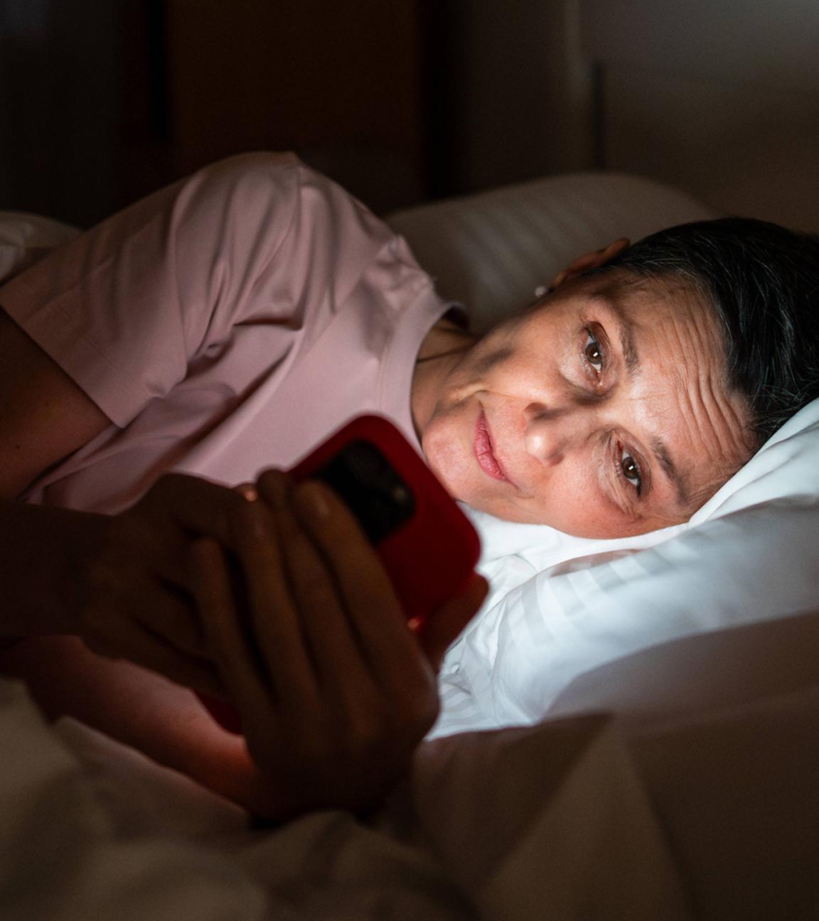Eine schläfrige, ältere Frau blickt abends in ihrem Bett auf ihr Smartphone.