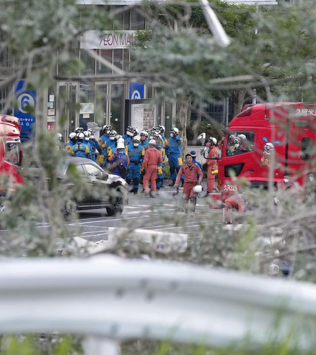 Ein Gruppe Rettungshelfer:innen vor der Aeon Mall in Japan, fotografiert durch ein Gebüsch.