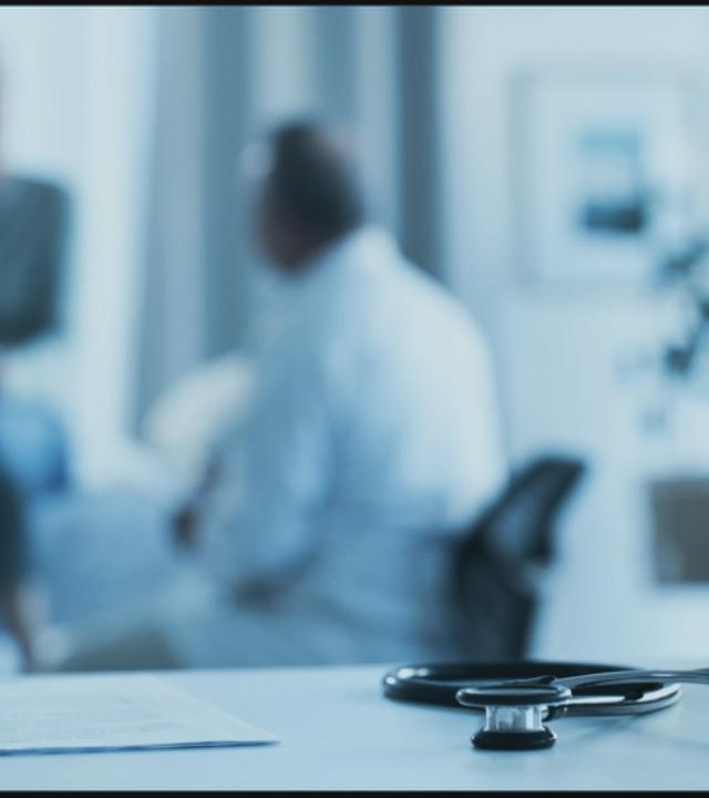 Stethoskop liegt auf Schreibtisch. Arzt mit Patient unscharf im Hintergrund zu sehen.