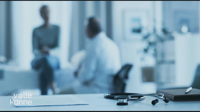 Stethoskop liegt auf Schreibtisch. Arzt mit Patient unscharf im Hintergrund zu sehen.