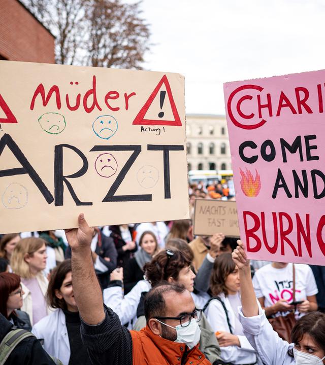 Ärzte stehen bei einem eintägigen Warnstreik des Marburger Bunds an der Charité mit einem Schild mit der Aufschrift "müder Arzt" und "Charite come in and Burnout".