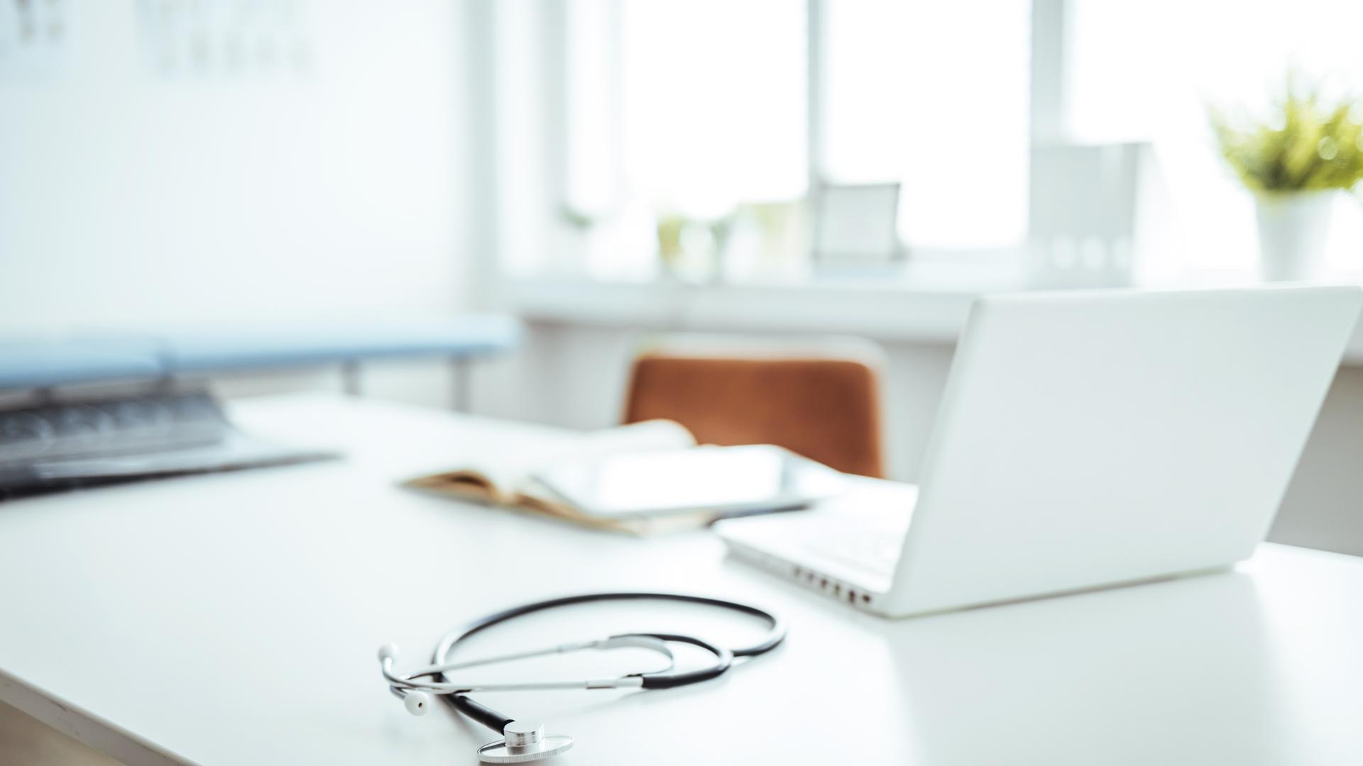 Symbolbild: ein Tisch mit Laptop und Stethoskop in einem Raum, der wie eine Praxis wirkt. 