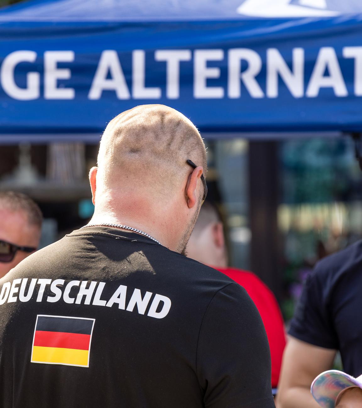 Rückansicht eines kahl rasierten Mannes mit der Aufschrift Deutschland auf seinem T-Shirt vor einem Stand der Jungen Alternativen.