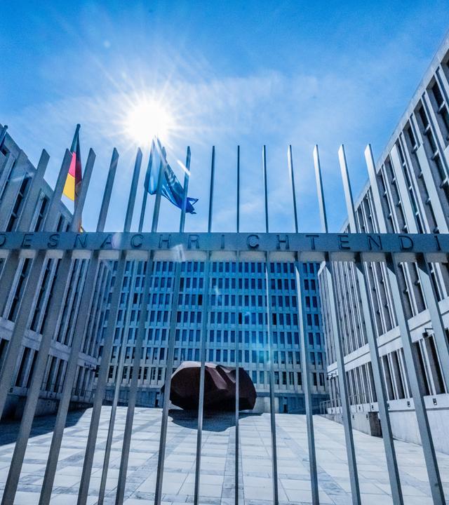 Das Tor des Bundesnachrichtendienstes ist zu sehen und das Gebäude im Hintergrund. Im Hof wehen die Deutschland- und die Europafahne.