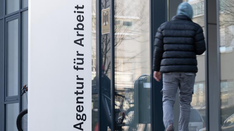 Ein Mann neben einer gläsernen Fassade hinter dem Schriftzug "Agentur für Arbeit".