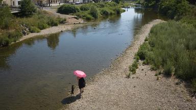 Eine Frau geht bei heißem Wetter im Ahrtal am Ufer der Ahr mit einem Hund spazieren.