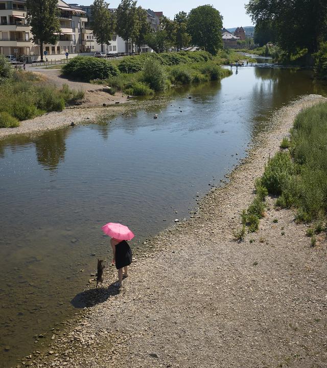 Eine Frau geht bei heißem Wetter im Ahrtal am Ufer der Ahr mit einem Hund spazieren.