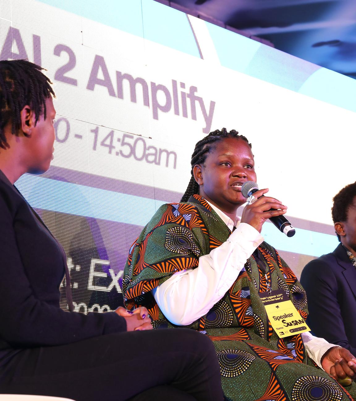 Auf einem Podium sitzten mehrere schwarze Personen. Eine Frau in buntem afrikanischem Gewand spricht in ein Mikrofon. Im Hintergrund wird der Schriftzug AI 2 Amplify eingeblendet.
