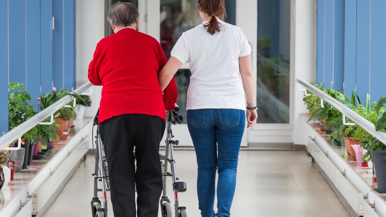pflegerin fuehrt seniorin mit rollator durch einen flur