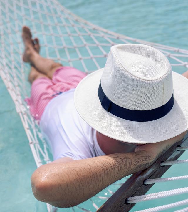 Ein junger Mann in Sommerkleidung und mit Hut liegt in einer Hängematte im klaren Meerwasser