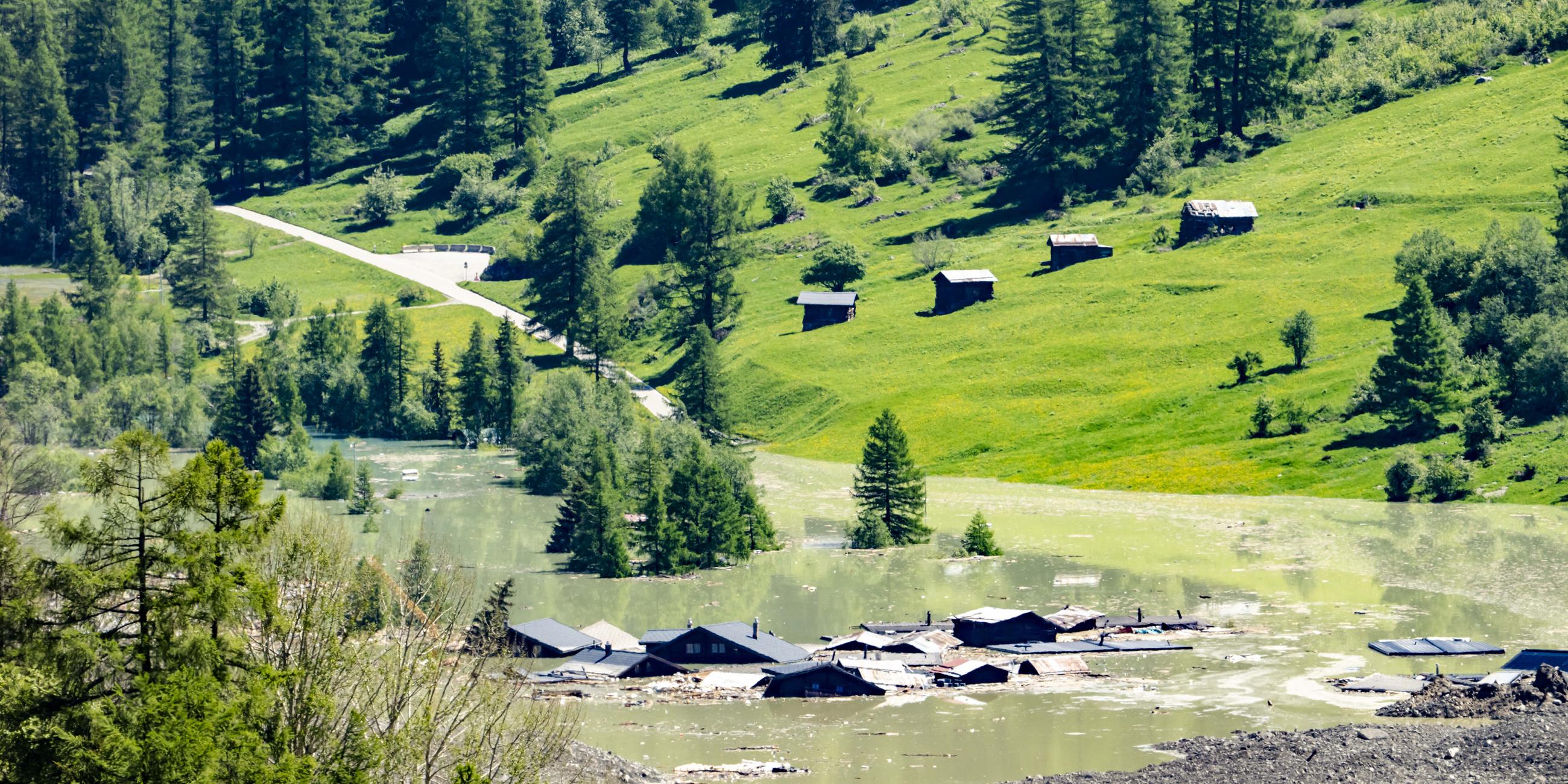30.05.2025, Schweiz, Blatten: Wasser aus dem Fluss Lonza fließt über Schlamm und Stein, nachdem sich ein See um den letzten Häusern des Dorfes Blatten gebildet hat.