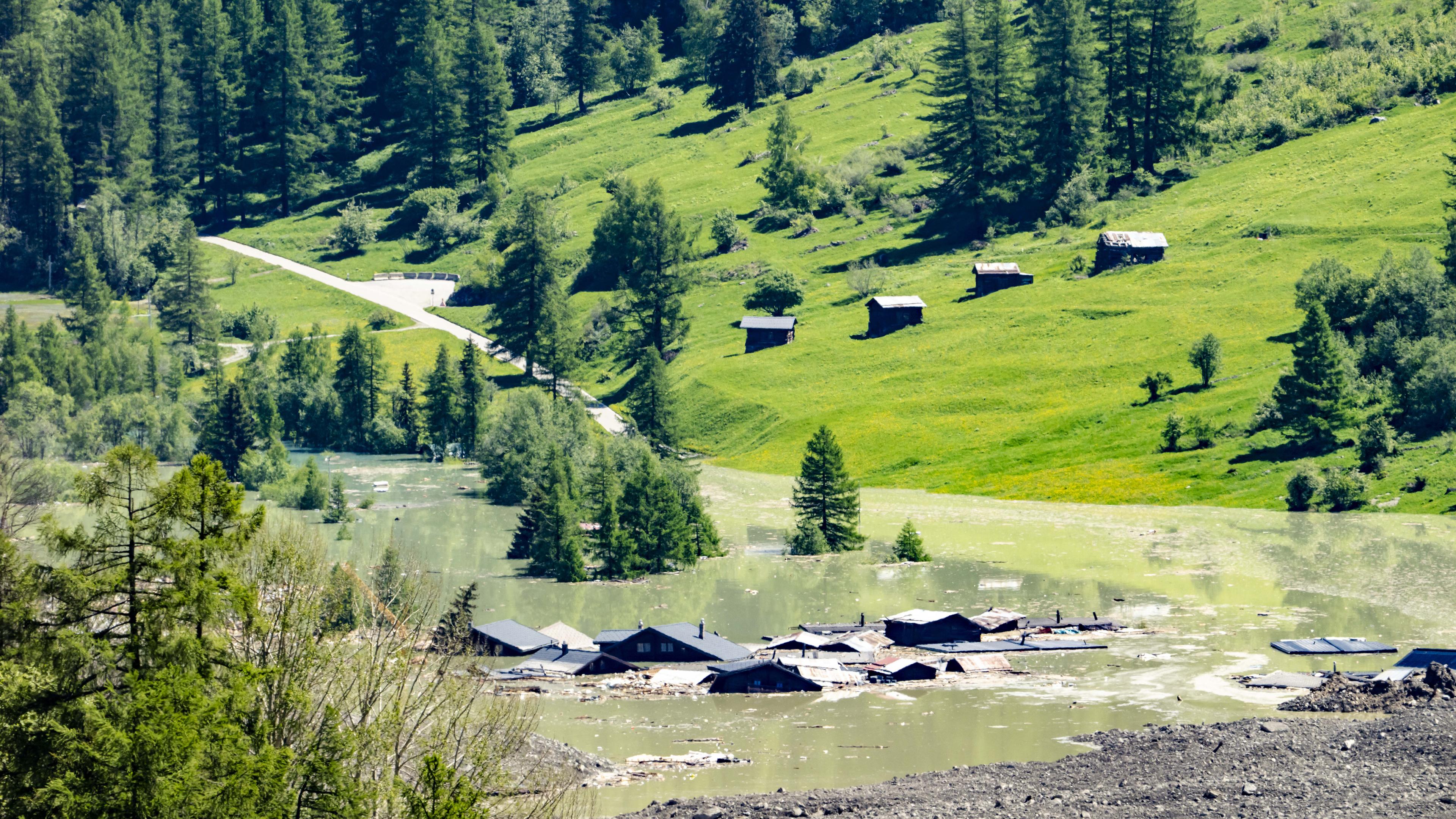 30.05.2025, Schweiz, Blatten: Wasser aus dem Fluss Lonza fließt über Schlamm und Stein, nachdem sich ein See um den letzten Häusern des Dorfes Blatten gebildet hat.