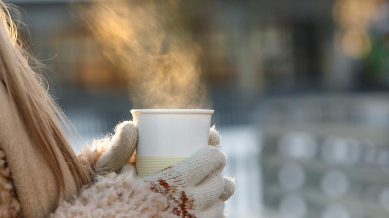 Frau mit Wollhandschuhen, die eine Tasse mit heißem Tee oder Kaffee im Winter hält, heißer Dampf kommt aus der Tasse