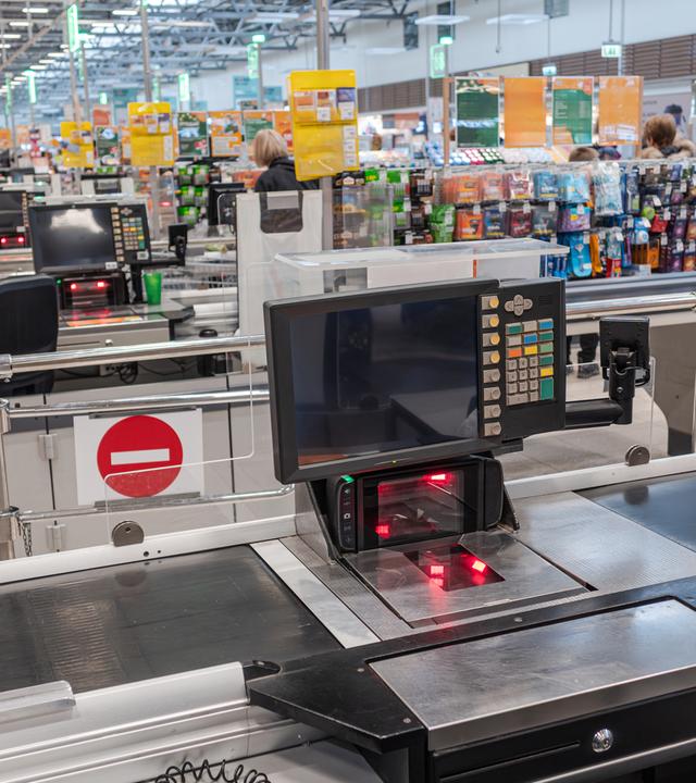 Das Bild zeigt einen leeren Supermarkt, im Vordergrund ist eine Kasse zu sehen.