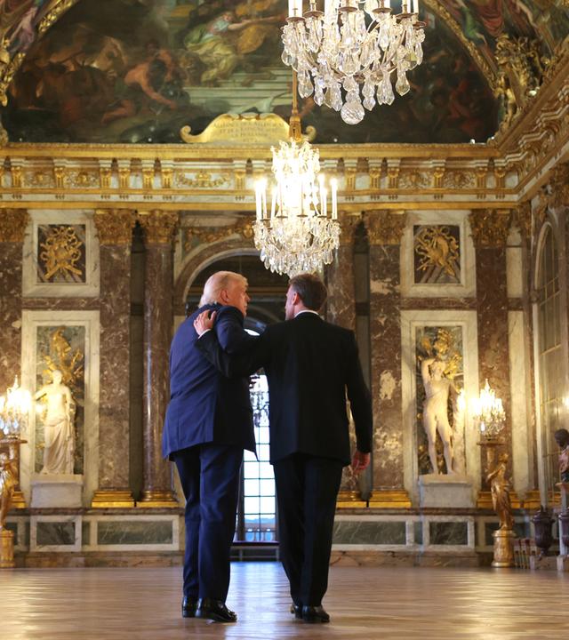 Donald Trump und Emmanual Macron laufen durch das Schloss Versailles. Macron hält seinen Arm über Trump.