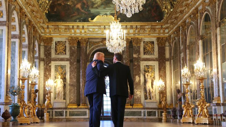 Donald Trump und Emmanual Macron laufen durch das Schloss Versailles. Macron hält seinen Arm über Trump.