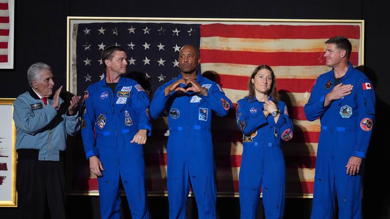 Harrison "Jack" Schmitt (von links) sowie die Artemis-II-Besatzungsmitglieder Reid Wiseman, Victor Glover, Christina Koch und Jeremy Hansen stehen auf der Bühne vor der US-Flagge. 