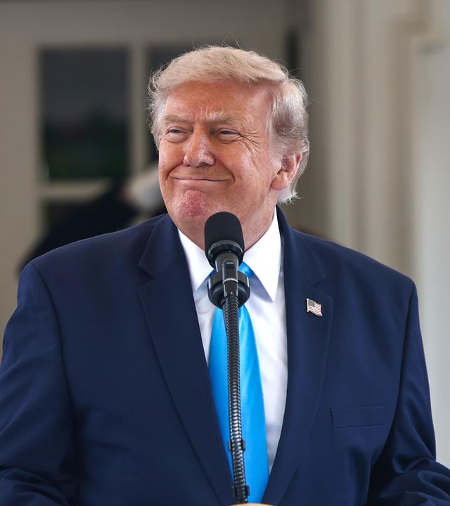 US-Präsident Trump steht vor einem Mikrofon und grinst.