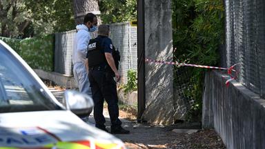 Ein Polizist in Uniform und ein Ermittler in weißem Untersuchungs-Anzug stehen vor einer Öffnung in einem Zaun, das mit Absperrband gebunden ist.