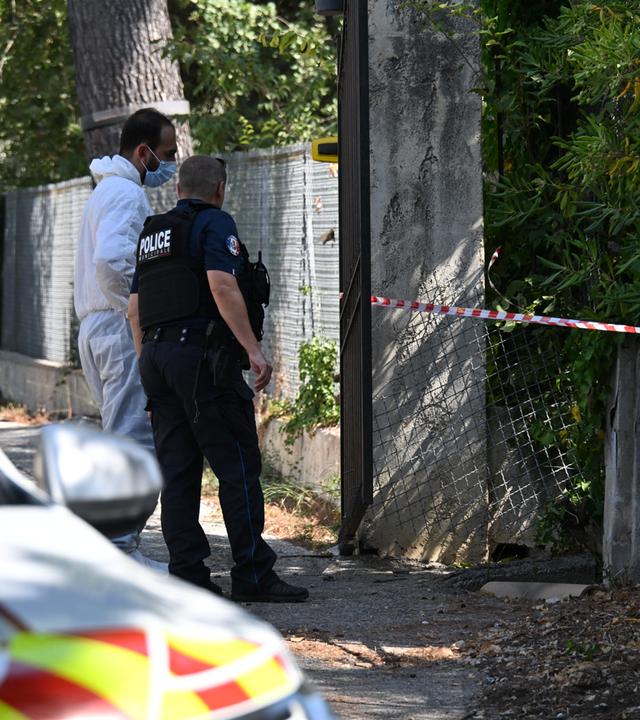 Ein Polizist in Uniform und ein Ermittler in weißem Untersuchungs-Anzug stehen vor einer Öffnung in einem Zaun, das mit Absperrband gebunden ist.
