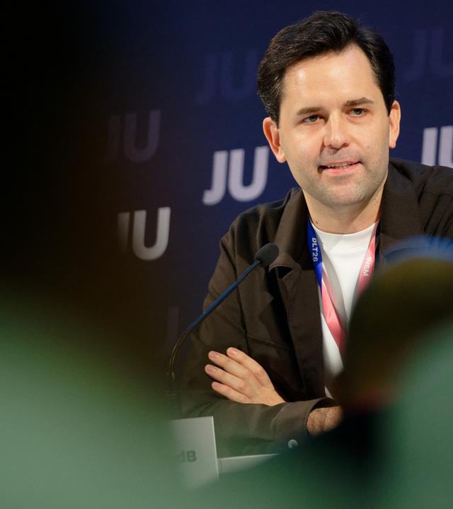 Johannes Winkel sitzt mit verschränkten Armen am Tisch vor einer Wand mit JU-Kürzeln.