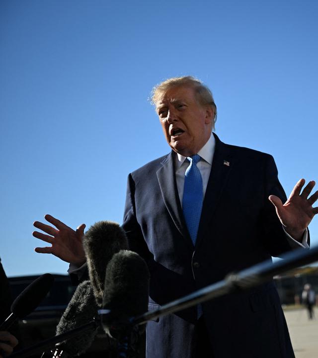 Trump steht vor Mikrofonen und wedelt ablehnend mit seinen Händen.
