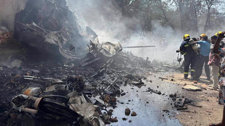 Absturzstelle des Flugzeugs an einem Haus, Verwüstung und Rauch, Feuerwehrleute stehen am rechten Bildrand.