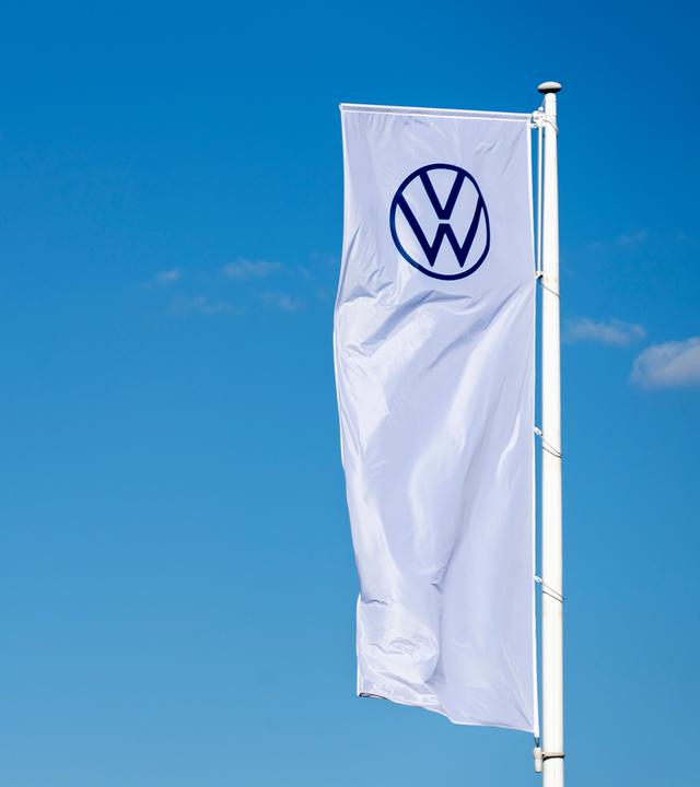 Rechts flattert eine lange weiße Flagge mit dem VW-Zeichen vor einem kräftig blauen Himmel. Links ragt eine orangefarbene, geometrische Form in das ansonsten freie Bild. Die Komposition begleitet das Thema eines möglichen europäischen Automobilherstellers nach Airbus-Vorbild, der zum Fortbestand der Marke Volkswagen beitragen soll.