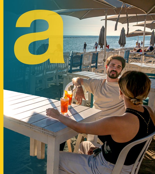 Ein Pärchen sitzt an Tisch am Strand in Italien, im Hintergrund Sonnenliegen, Meer und Touristen
