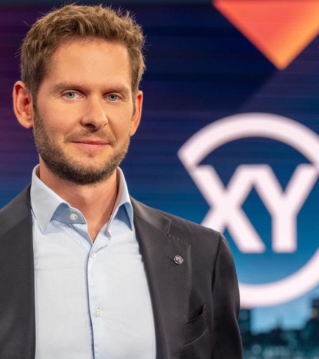 Ein mittelalter Mann in dunkelgrauem Anzug schaut in die Kamera. Im Hintergrund ist ein großes XY zu sehen.