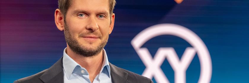 Ein mittelalter Mann in dunkelgrauem Anzug schaut in die Kamera. Im Hintergrund ist ein großes XY zu sehen.