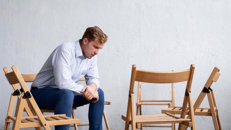 Ein trauriger Mann sitzt in gebeugter Haltung in einer Gruppe aus leeren Stühlen.