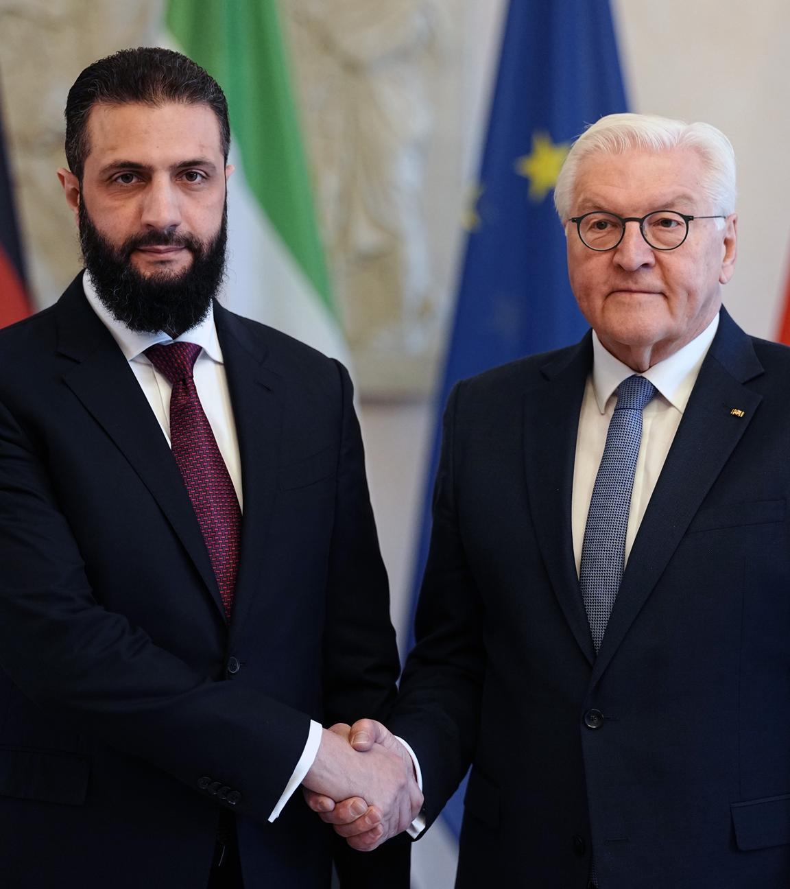 Syriens Übergangspräsident Ahmed al-Scharaa und Bundespräsident Frank-Walter Steinmeier schütteln die Hände, beide mit ausdruckslosem Gesicht vor den Flaggen Deutschlands, Syriens und der EU.